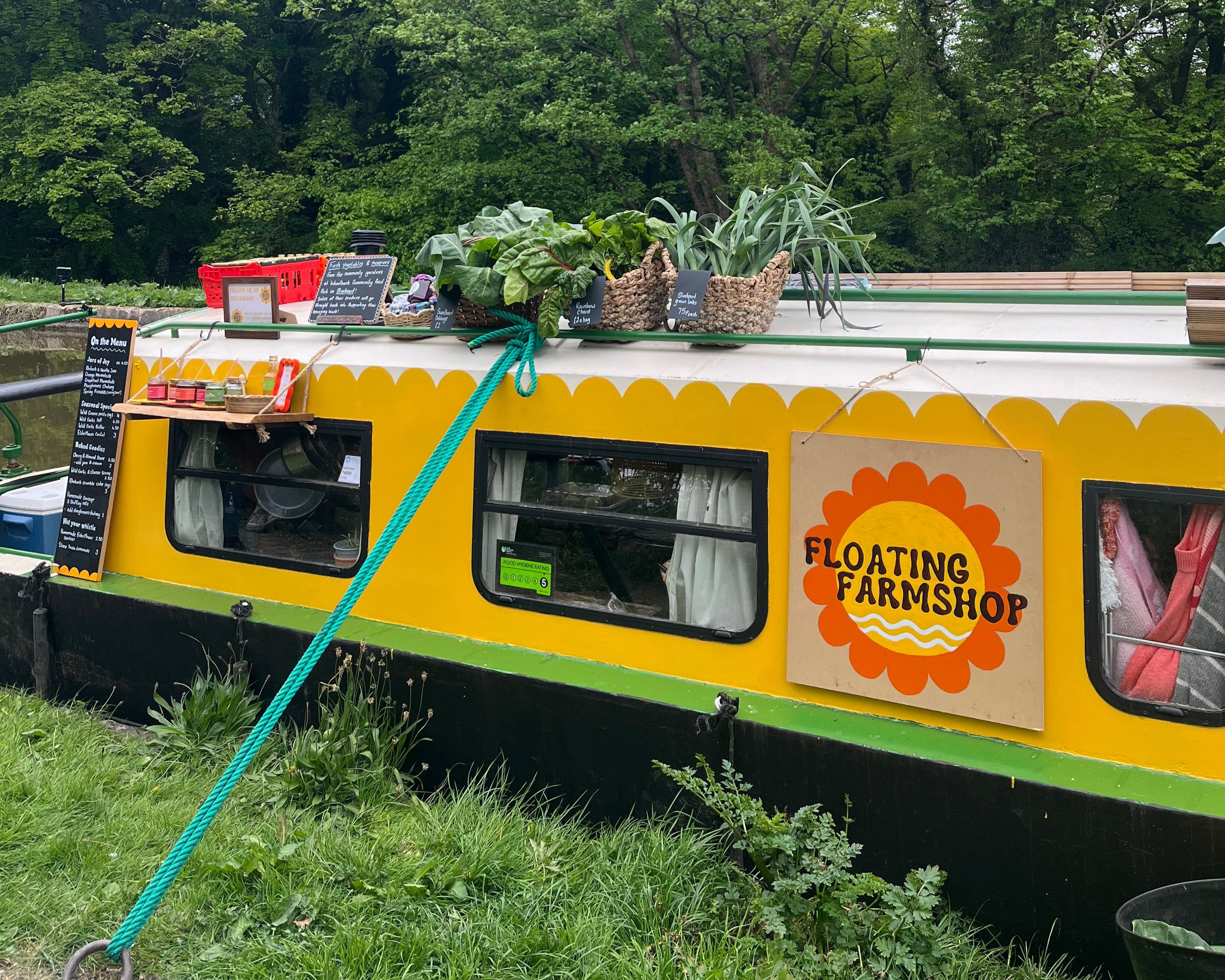 Yellow floating farmshop with 'Floating Farmshop' branding on a canal surrounded by greenery.